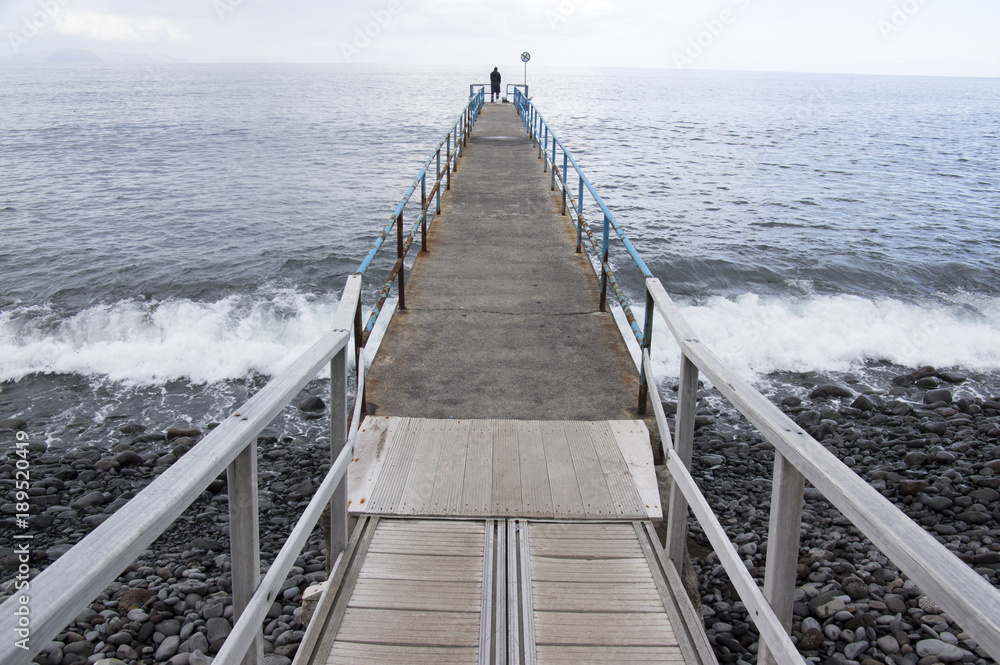 Fototapeta premium Wooden port pier with fisherman silhouette on the end, wild ocean with waves, Funchal, Madeira, Portugal