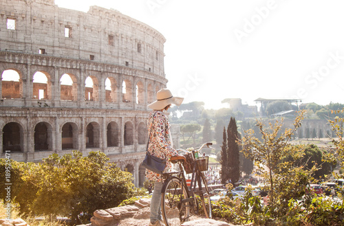 Canvas Print Beautiful young woman in colorful fashion dress walking alone on hill with her bike in front of colosseum in Rome at sunset with trees