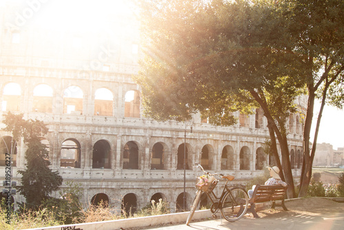 Photography Beautiful young woman in colorful fashion dress sitting alone on a bench with bike looking at colosseum in Rome at sunset under a tree