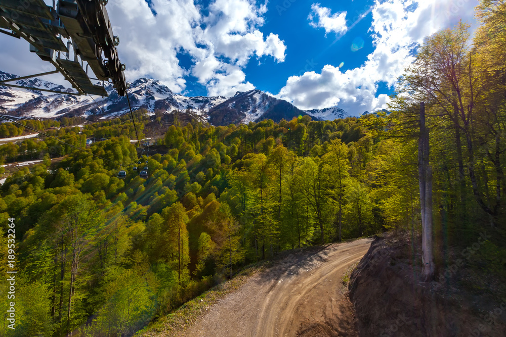 The cable car leads to the top of the snow-capped mountain above the green trees of the forest and the dirt road below. Spring mountain landscape, Sochi, Russia.