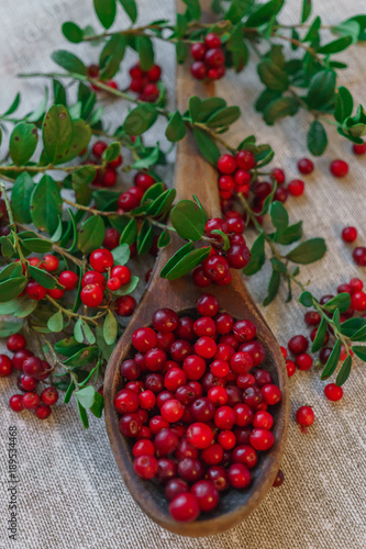 Berries of red lingonberry in a wooden spoon, along with twigs, on a table.