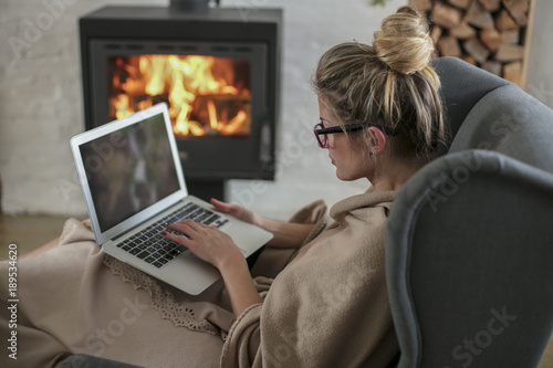 Beautiful middle-aged woman next to the fireplace relaxes in the living room and works on laptop from house. Girl in 30's surveyed for the  home bussiness project