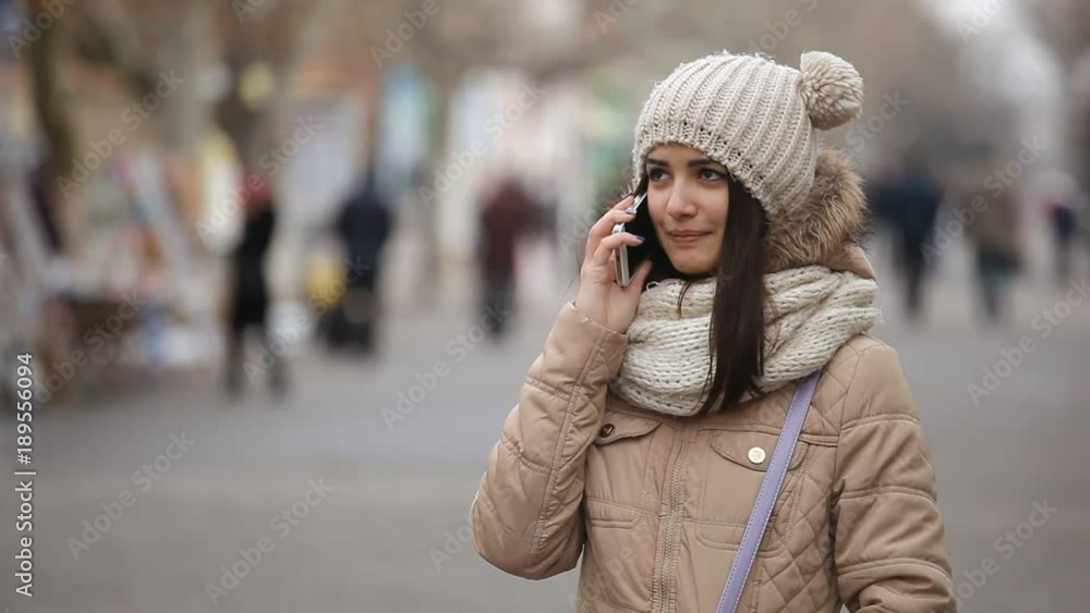 An astonishing view of a gorgeous girl in a knitted hat with cheery balls who smiles and speaks on her smart phone on an arty alley in winter An astonishing view of a gorgeous girl in a knitted hat with cheery balls who smiles and speaks on her smart phone on an arty alley in winter