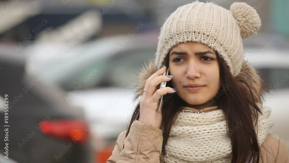 A wonderful view of a cheery young woman in a knitted hat with balls, a fur anorak, calling by her mobile near a white brand new car. A wonderful view of a cheery young woman in a knitted hat with balls, a fur anorak, calling by her mobile near a white brand new car.