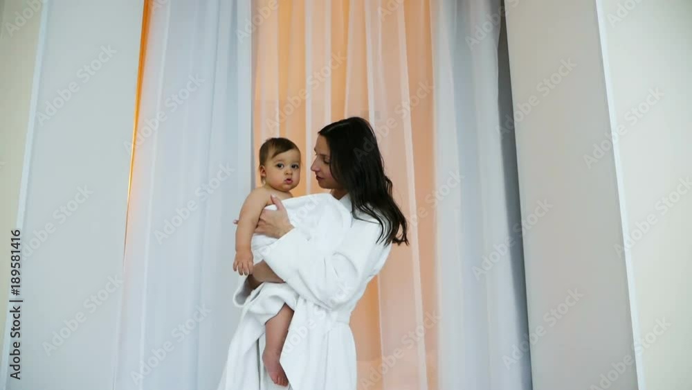woman in white Bathrobe holding son in her arms in bright bedroom woman in white Bathrobe holding son in her arms in bright bedroom