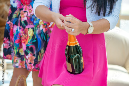  Girl in pink dress is opening champagne bottle to pour the drink in glasses. Cork is about to pop out. Cheerful bride and bridesmaids party