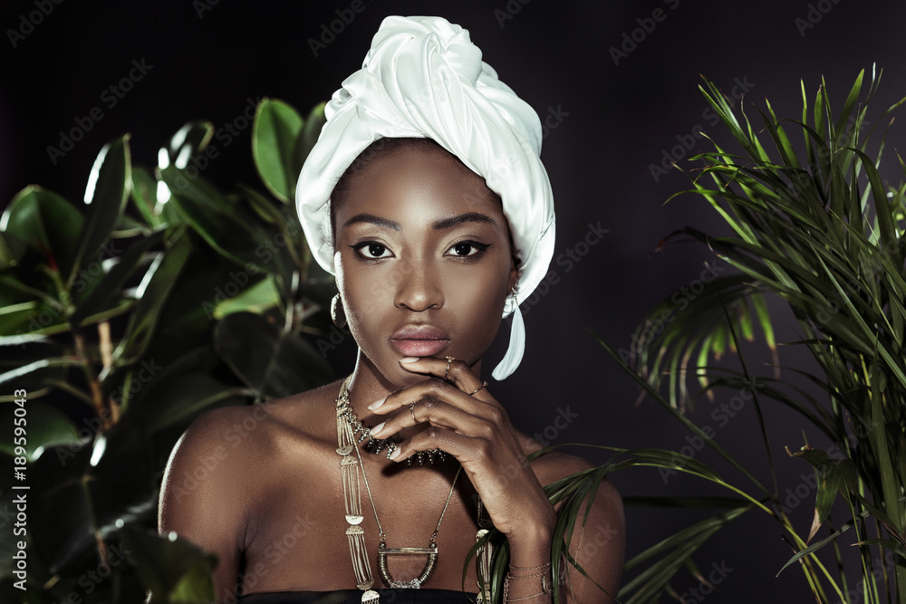 young african american woman in white wire head wrap behind leaves looking at camera young african american woman in white wire head wrap behind leaves looking at camera