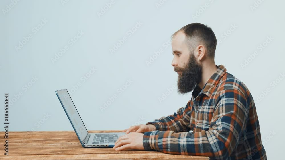 Bearded hipster man sitting at table working with laptop, turning to camera and smiling side view Bearded hipster man sitting at table working with laptop, turning to camera and smiling side view