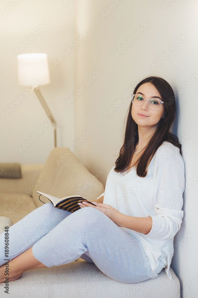 Photo of smart female student does test writing in notepad, prepares for examination while sits at comfortable sofa, wears glasses and casual clothes. Talented person works on creating design interior Photo of smart female student does test writing in notepad, prepares for examination while sits at comfortable sofa, wears glasses and casual clothes. Talented person works on creating design interior