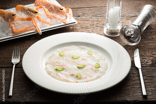 Scallops carpaccio served with langoustines on a white plate and wooden rustic brown  table with dinner settings. Close up 
