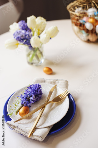 easter holiday dining table with golden metallic cutlery and eggs, hyacinth and tulip bouquet, blue and white plates. Celebration concept