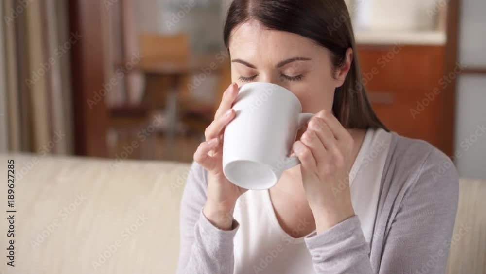 Young woman relaxing sitting on couch at home in the morning. Pretty female drinking tasty coffee or tea from white cup in living room