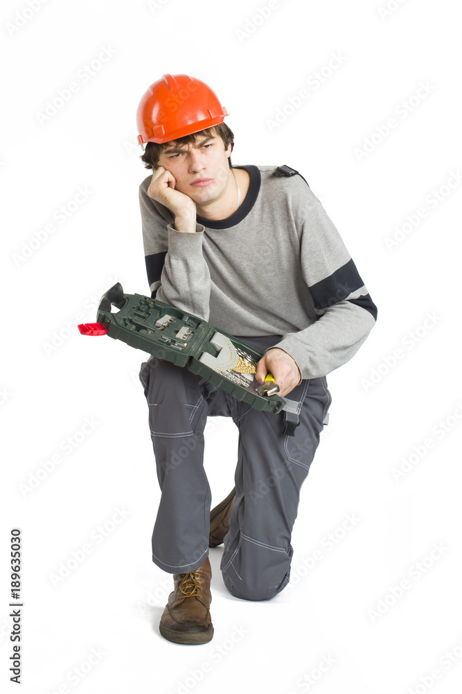 A young man in working grey clothes and orange hard helmet sitting with toolkit on isolated white background. A young man in working grey clothes and orange hard helmet sitting with toolkit on isolated white background.