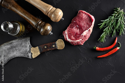 Piece of beef tenderloin, with ax for cutting and chopping meat, spices were cooking - rosemary, pepper on old black table. Top view copy space photo