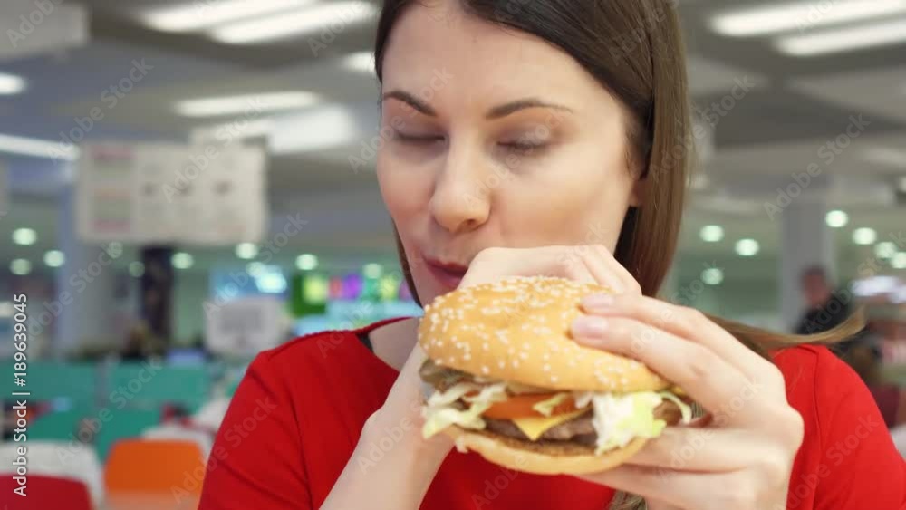Hungry girl eating hamburger on food court. Woman biting cheeseburger at fast food restaurant Hungry girl eating hamburger on food court. Woman biting cheeseburger at fast food restaurant