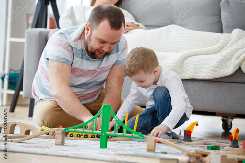 Picture of father playing with son in toys sitting on floor, sitting next to pregnant mother
