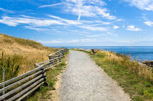 Empty Gravel Coastal Path with a Wooden Bench Facing the Ocean on a Sunny Summer Day.