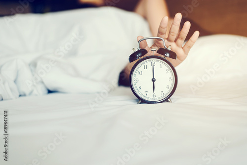 The woman's hand under the blanket reaches out for the alarm clock.