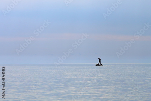Silhouette of stand up paddle boarder paddling on a flat warm quiet sea. Water tourist with hermetic bags on SUP