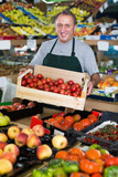 Positive male employee is standing with box with tomatos