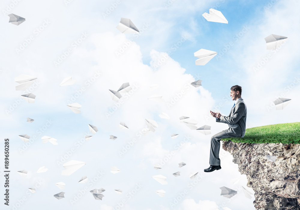 Elegant businessman on rock edge making calls and paper planes f Elegant businessman on rock edge making calls and paper planes f