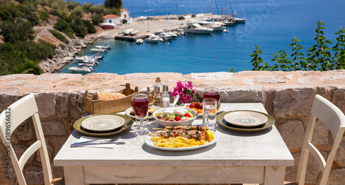 A table served for two with chicken souvlaki and french fries, greek salad, snacks and drinks on the summer terrace of the hotel room by the seascape, beautiful summer greek holidays concept.