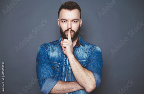 Handsome confident young bearded man in a denim shirt shows a finger more quietly on a gray background.