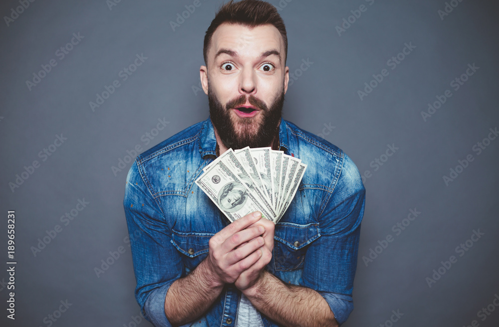 Monetary winnings. Young joyful man in a denim shirt holds a lot of dollars in the hands and shocked looking at the camera on a gray background. Monetary winnings. Young joyful man in a denim shirt holds a lot of dollars in the hands and shocked looking at the camera on a gray background.