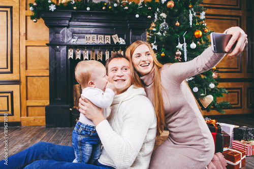 Christmas theme. young family with blond boy of one year sits on wooden floor against background of a Christmas tree with gifts and makes selfie, self-portrait on the front camera of a silver phone photo