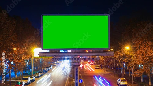 A billboard with green chroma key on a background of a city night landscape of fast moving cars with long exposure. Time Lapse video. The camera is approaching