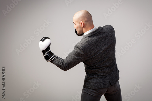 Bald man, confident manager in a white shirt, gray suit and boxing gloves makes an uppercut on a white isolated background, side view photo