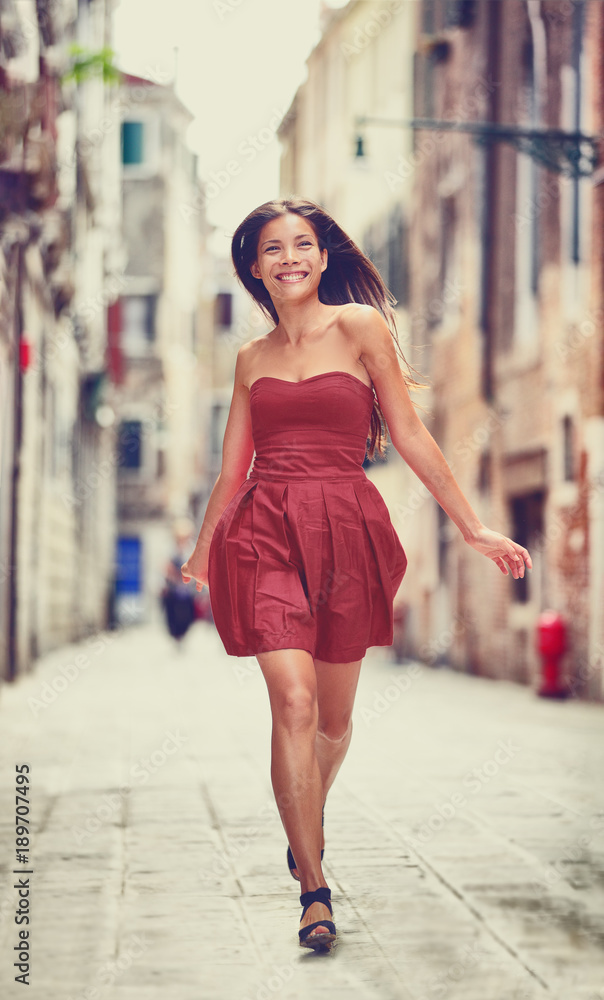 Happy beautiful woman in red summer dress walking and running joyful and cheerful smiling in Venice, Italy. Pretty sexy fashion model girl in her 20s. Mixed race Asian Caucasian female model outside.
