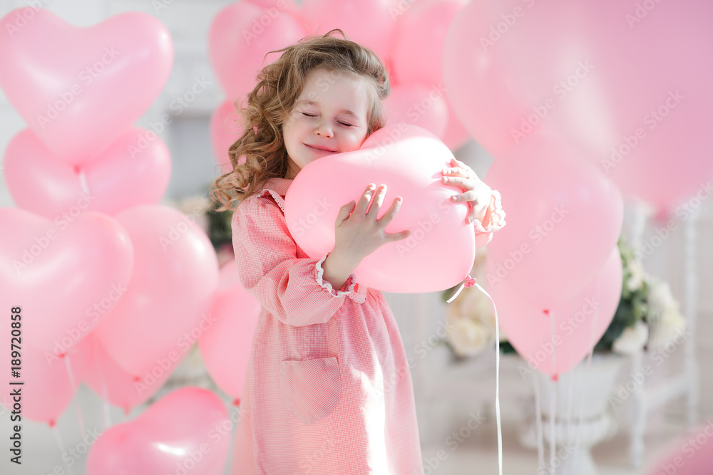 A little girl of 6 years with long curly hair, a beautiful smile, sits alone in a large bright room with lots of pink balloons in the shape of a heart. Valentine's Day and party celebration. A little girl of 6 years with long curly hair, a beautiful smile, sits alone in a large bright room with lots of pink balloons in the shape of a heart. Valentine's Day and party celebration.