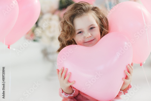 A little girl of 6 years with long curly hair,a beautiful smile, sits alone in a large bright room with lots of pink balloons in the shape of a heart. Valentine's Day and party celebration.