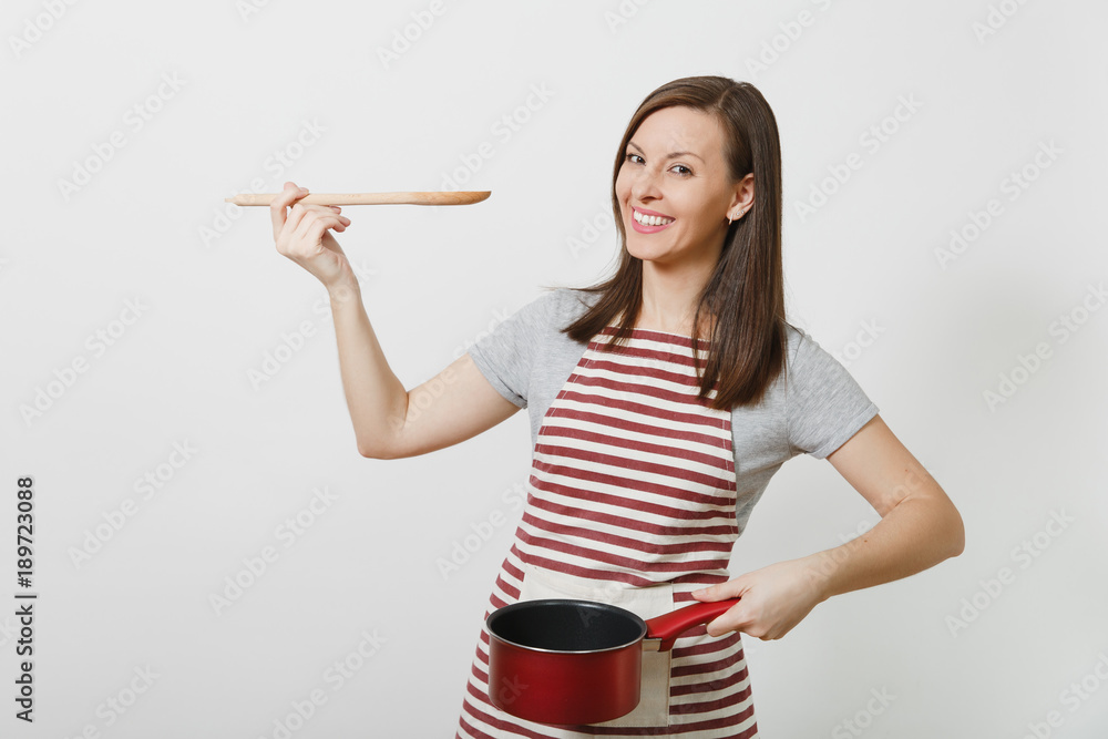 Young smiling caucasian housewife in striped apron, gray t-shirt isolated on white background. Beautiful housekeeper woman holding tasting red empty stewpan wooden spoon. Copy space for advertisement. Young smiling caucasian housewife in striped apron, gray t-shirt isolated on white background. Beautiful housekeeper woman holding tasting red empty stewpan wooden spoon. Copy space for advertisement.