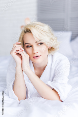 pensive attractive blonde woman in bathrobe looking away in bedroom