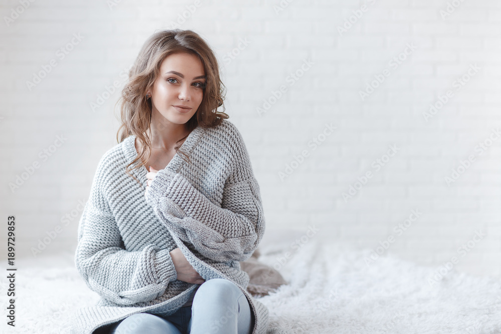 Cute young girl in a gray knitted sweater. Beautiful woman is relaxing in a  white bedroom.