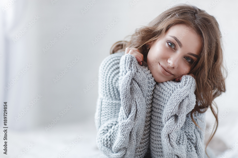 Fototapeta premium Cute young girl in a gray knitted sweater. Beautiful woman is relaxing in a white bedroom. Beautiful women in winter clothes are waking up in the morning. Woman wearing a sweater in a white bedroom.