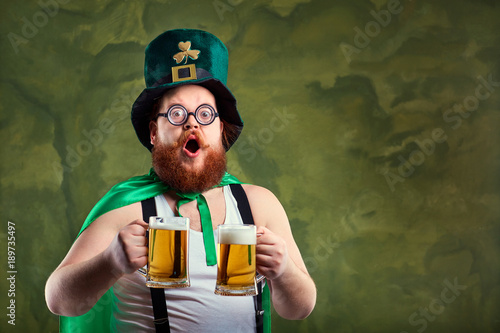 A fat man with a beard in St. Patrick's suit is smiling with a mug of beer on a green background. photo