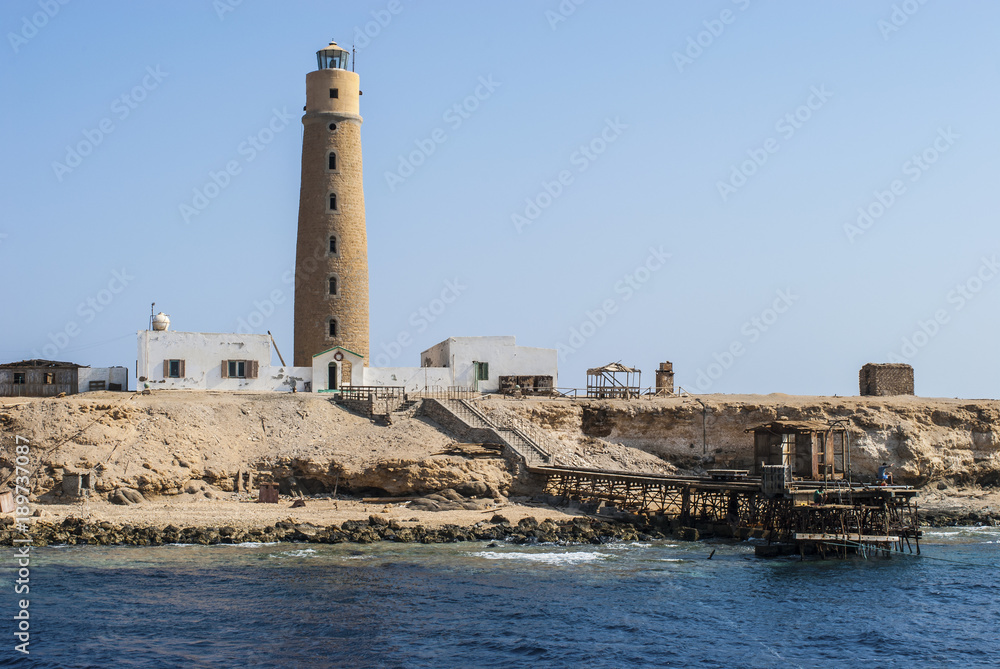 A scenic view of a seashore with an old picturesque lighthouse open for visitors, and a long, wooden pier stretching out into the sea. The bright landscape made from the Red Sea in Egypt.