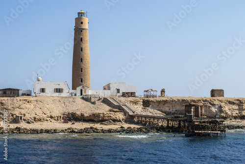 A scenic view of a seashore with an old picturesque lighthouse open for visitors, and a long, wooden pier stretching out into the sea. The bright landscape made from the Red Sea in Egypt.