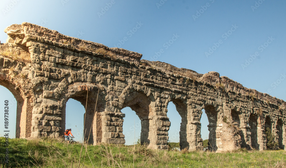 Cyclist riding bike on dirt road under ancient roman aqueduct in a park. Young attractive athletic man with orange sportswear and backpack in parco degli acquedotti in Rome Cyclist riding bike on dirt road under ancient roman aqueduct in a park. Young attractive athletic man with orange sportswear and backpack in parco degli acquedotti in Rome