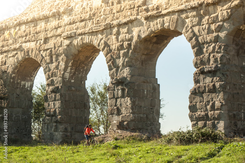 Canvas Print Cyclist riding bike on dirt road under ancient roman aqueduct in a park