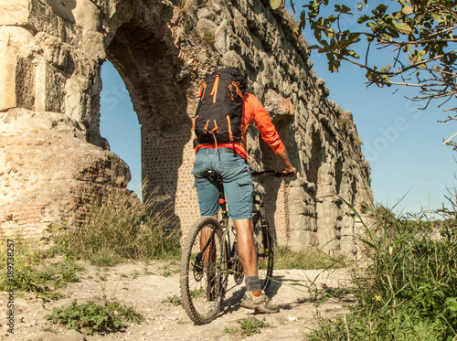 Photography Cyclist riding bike on dirt road under ancient roman aqueduct in a park