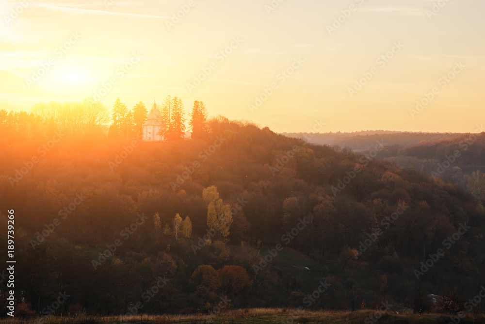 Fototapeta premium Landscape of old solitary church on the hill with autumn forest during the sunset