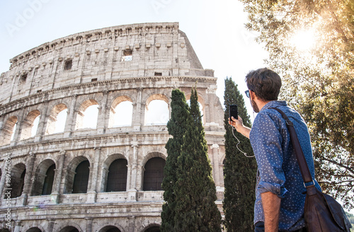 Happy young man tourist wearing blue shirt at colosseum using smartphone taking pictures in Rome on sunny day. Lens flare.