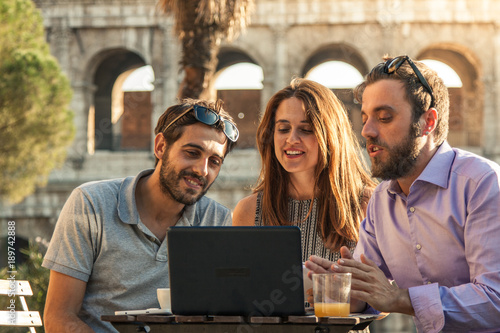 Photography Three happy friends using laptop having video call sitting at bar restaurant table in front of colosseum in rome at sunset