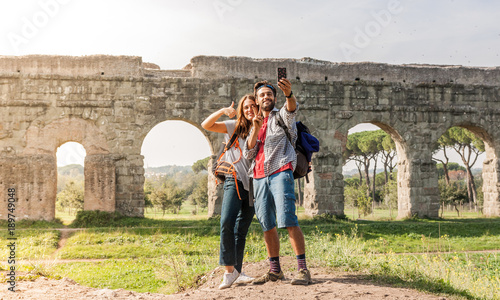 Fotografie Young couple backpackers tourists with guitar taking selfies in front of roman aqueduct ruins in acquedotti park in rome