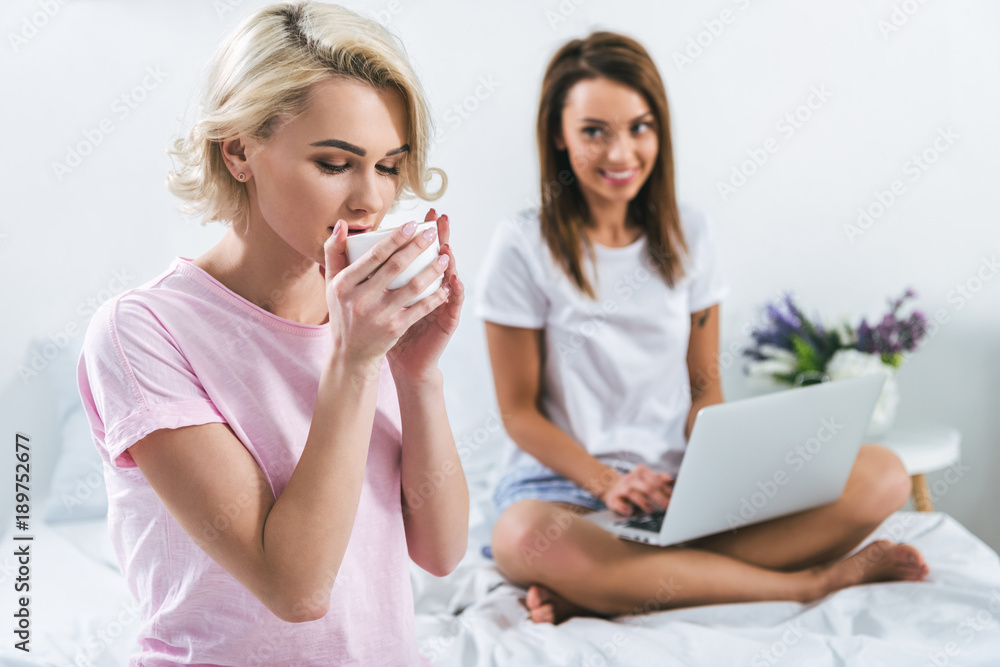 female friends drinking coffee and using laptop on bed female friends drinking coffee and using laptop on bed