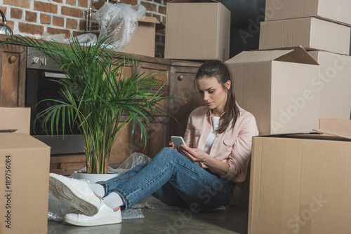 young woman using smartphone while sitting between cardboard boxes in new apartment